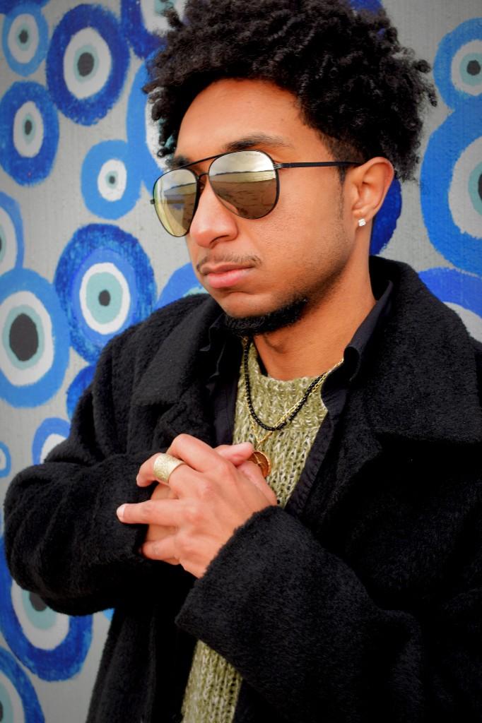Layered gold ring, necklaces, and stud earring with layered outerwear, against a blue patterned background.