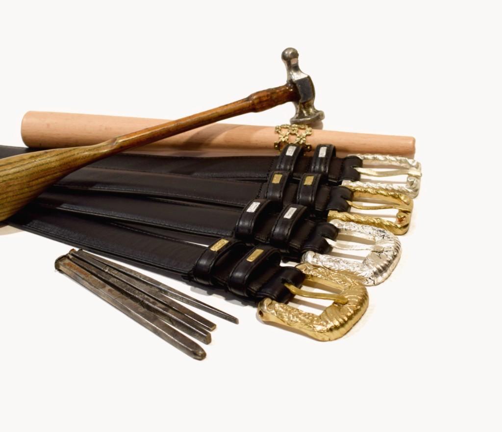 Engraved black leather belts with brass and silver buckles, hand tools, and bench metalwork on a white studio background.